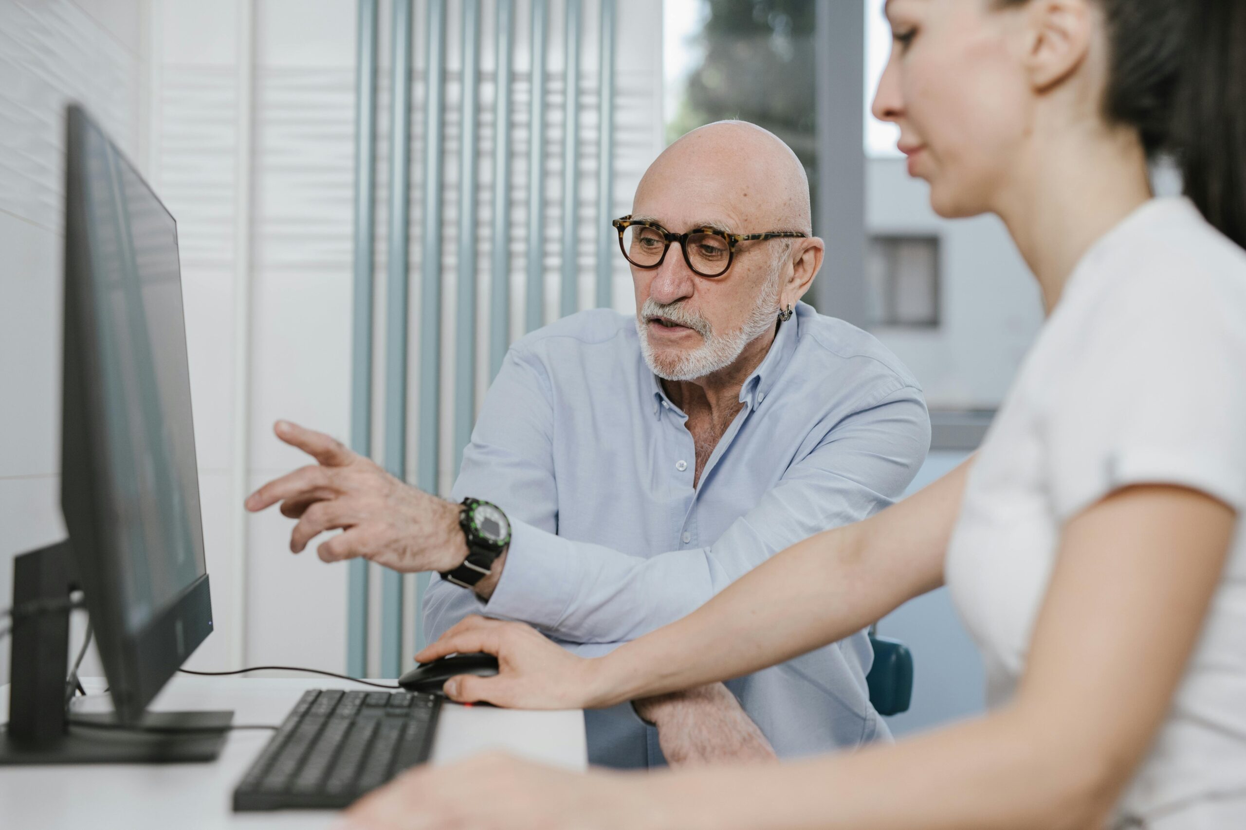 An elderly man pointing at a computer screen while assisting a woman in an office setting.